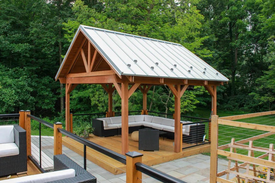 Covered wood pavilion with metal roof, deck railings, outdoor seating, and wooded backyard beyond.